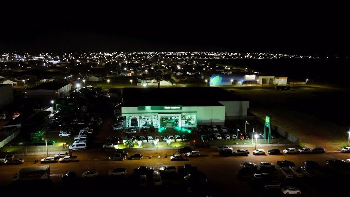 Vista aérea noturna da concessionária iluminada, com o pátio cheio